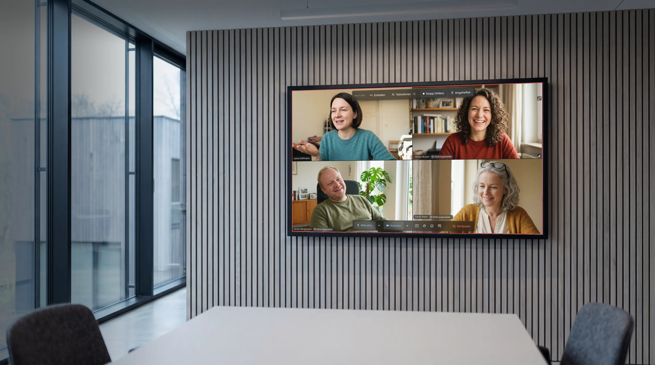 Besprechungsraum mit Glaswand und Eichenlamellen; auf dem Wandbildschirm läuft eine Raven-Videokonferenz mit vier Teilnehmenden