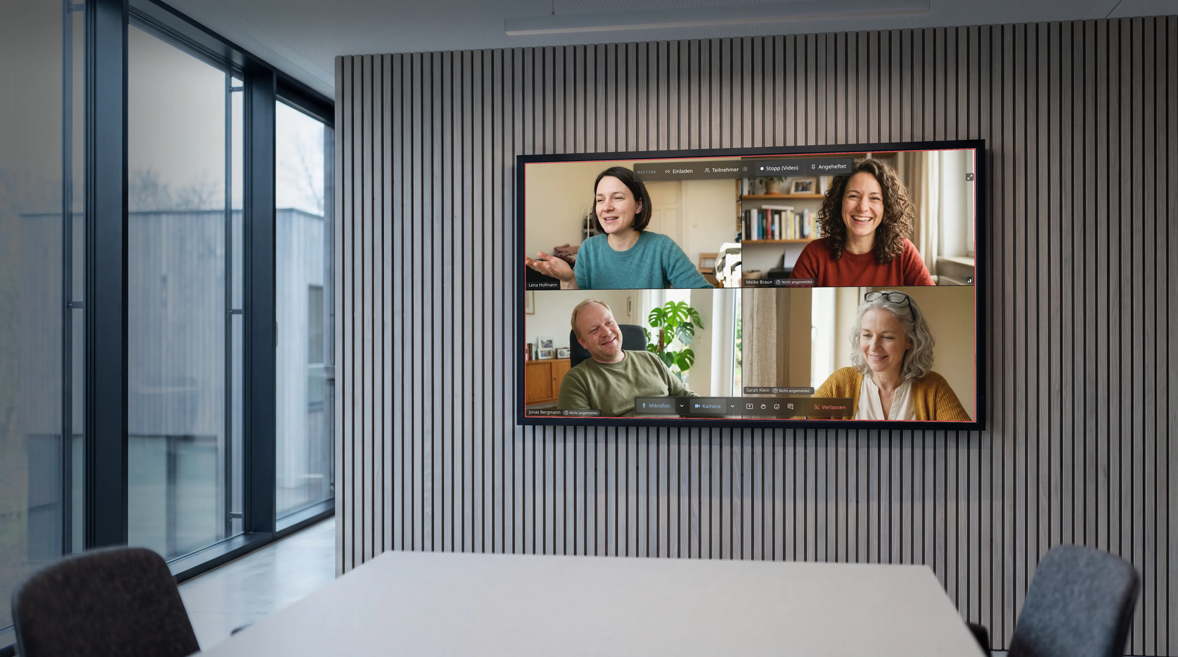 Besprechungsraum mit Glaswand und Eichenlamellen; auf dem Wandbildschirm läuft eine Raven-Videokonferenz mit vier Teilnehmenden