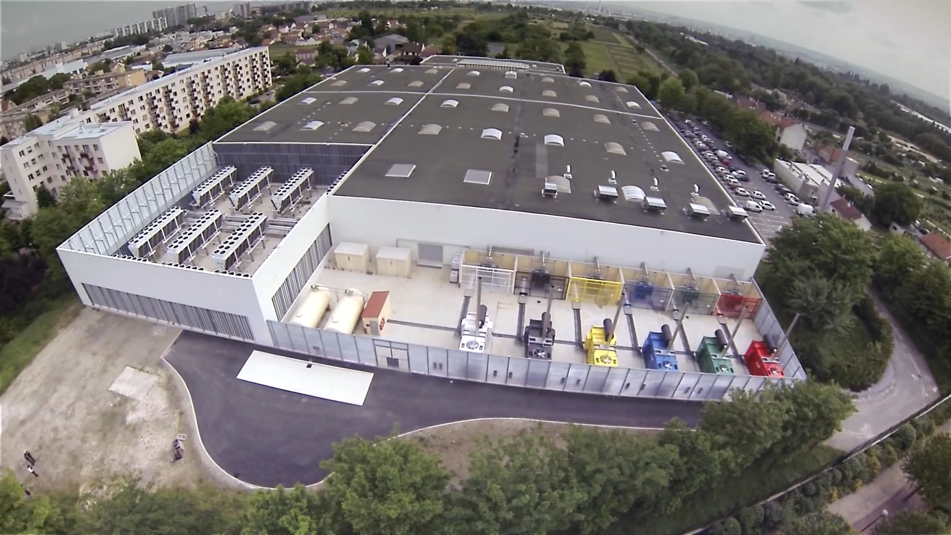 Luftbild des Rechenzentrums DC3 in Vitry-sur-Seine bei Paris: eine flache weiße Halle mit Kühlaggregaten auf dem Dach, davor eine Reihe farbiger Notstromaggregate hinter einem Zaun